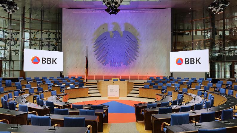Frontalblick auf den alten Bundestag in Bonn. Präsent ist ein großer Bundesadler, ein großer Teppich mit einem Zivilschutzzeichen sowie links und rechts zwei Bildschirme auf denen die Abkürzung BBK steht.
